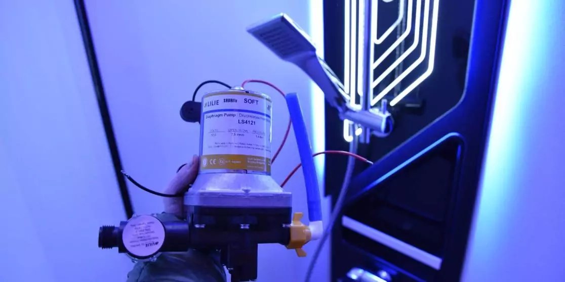 Hand mit Schutzhandschuh h&auml;lt ein zylindrisches Ger&auml;t mit Etikett; links ein runder Manometer; daran angeschlossene Schl&auml;uche und Kabel; im Hintergrund ein vertikales, beleuchtetes Paneel mit wei&szlig;en Linien und blauem Licht