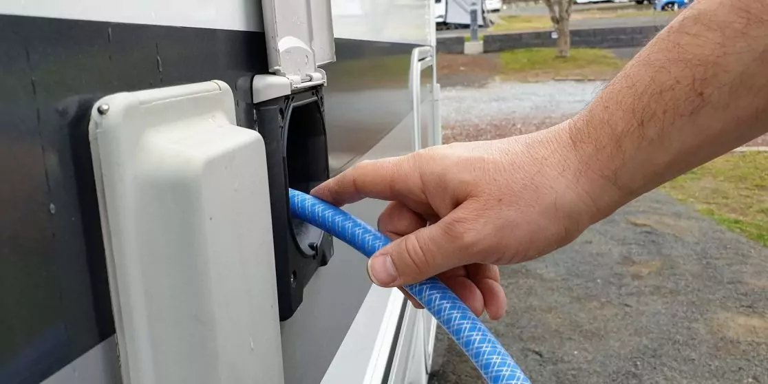 Hand f&uuml;hrt blauen flexiblen Schlauch in rechteckigen Anschluss an der Au&szlig;enseite eines Wohnwagens; neben dem Anschluss eine wei&szlig;e Abdeckung; im Hintergrund Parkfl&auml;che mit Gras und B&auml;umen