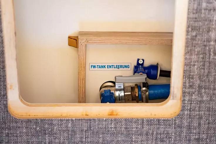 Rechteckige &Ouml;ffnung in einer Wand mit sichtbaren blauen Rohrleitungen, Metallschellen, blauem Absperrhahn und einem Aufkleber mit der Aufschrift "FW-TANK ENTL&Uuml;FTUNG"