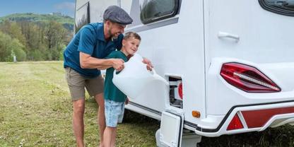 Mann und Junge gie&szlig;en Wasser aus einem wei&szlig;en Kanister in das Einf&uuml;llfach eines Wohnwagens auf einer Wiese mit H&uuml;geln im Hintergrund