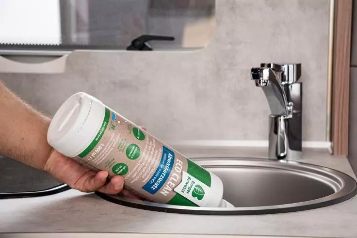 Hand h&auml;lt eine wei&szlig;e Plastikdose mit gr&uuml;nem Etikett &uuml;ber ein rundes Edelstahl-Sp&uuml;lbecken, daneben ein verchromter Wasserhahn auf einer Arbeitsplatte