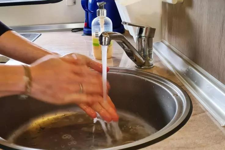 Zwei H&auml;nde unter flie&szlig;endem Wasser aus einem Wasserhahn in ein Edelstahlsp&uuml;lbecken; Seifenspender und Arbeitsplatte im Hintergrund sichtbar