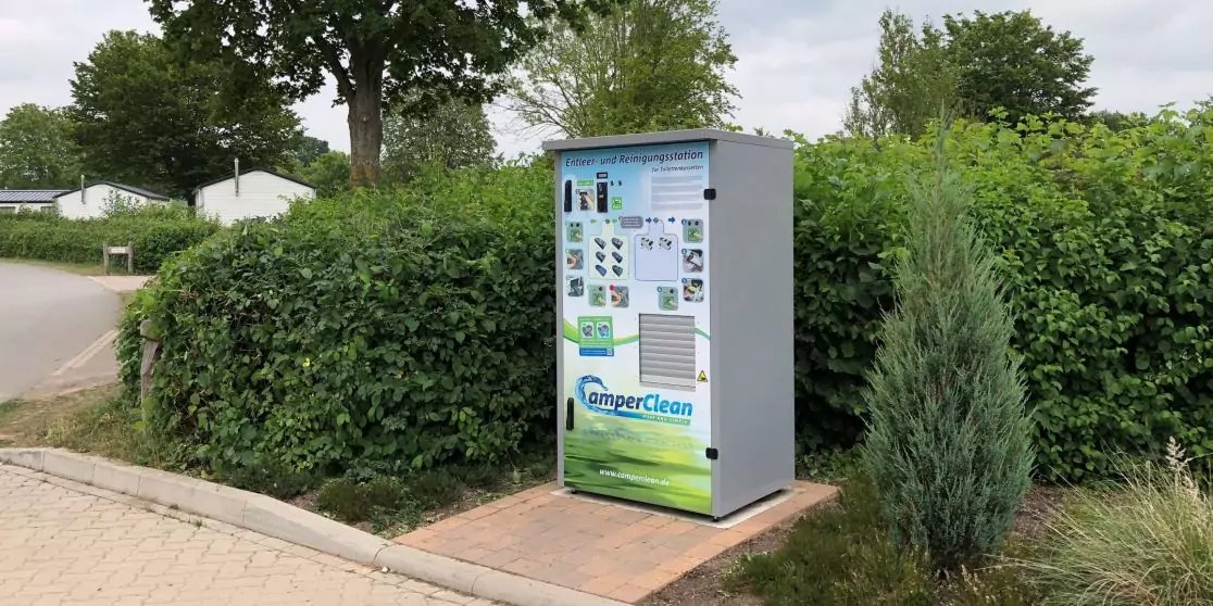 Rechteckiger metallener Schrank mit Aufklebern und der Aufschrift "CamperClean" auf einem gepflasterten Bereich neben einer Hecke und einem kleinen Nadelbaum, im Hintergrund H&auml;user, B&auml;ume und bew&ouml;lkter Himmel