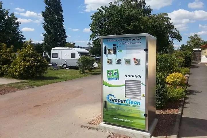 wei&szlig;e Service-S&auml;ule mit Aufschrift "CamperClean" im Vordergrund; im Hintergrund Wohnwagen und Pkw, B&auml;ume, Str&auml;ucher, Gehweg und blauer Himmel mit Wolken