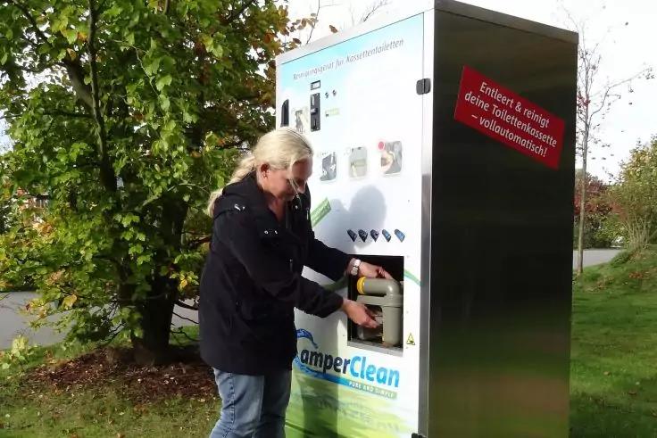 Person mit hellen Haaren steckt einen Gegenstand in einen Automaten mit der Aufschrift "CamperClean" im Freien; links B&auml;ume, rechts Wiese