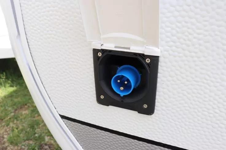 Blauer runder Steckdoseneinsatz mit drei Metallstiften in schwarzer quadratischer Einfassung an wei&szlig;er strukturierter Au&szlig;enwand, ge&ouml;ffnete wei&szlig;e Abdeckung dar&uuml;ber