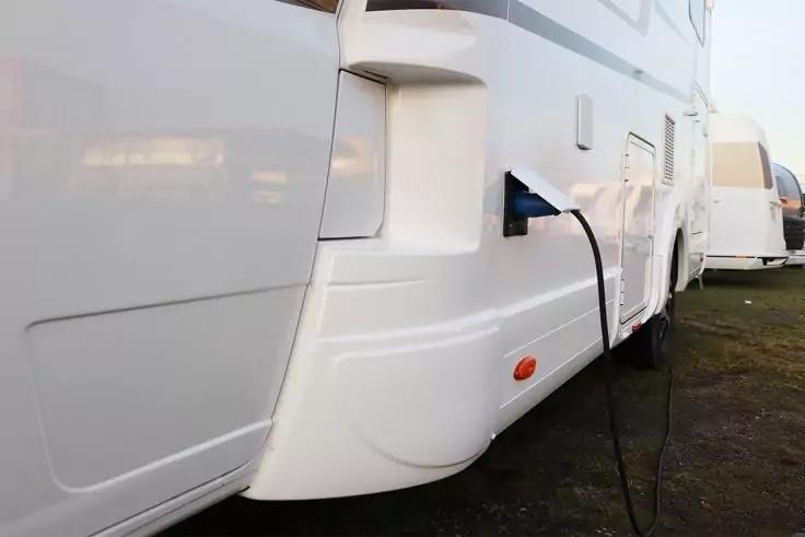 Wei&szlig;es Wohnmobil auf Gras, ein Stromkabel in eine Seitensteckdose eingesteckt, im Hintergrund weiteres Wohnmobil