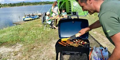 Person wendet Fleischst&uuml;cke mit Zange auf einem Grill am Ufer; im Hintergrund Zelt, Tisch mit Personen und Boote auf dem See