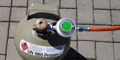 Propangasflasche mit angeschlossenem Druckregler und orangefarbenem Schlauch auf gepflastertem Untergrund; Aufkleber 'UN 1965 Propan' und Gefahrensymbol
