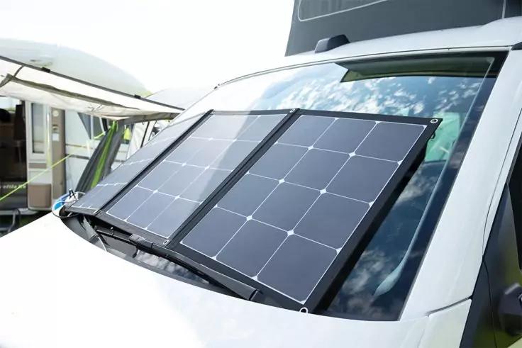 Faltbares Solarpanel mit zwei schwarzen Paneelen auf der Windschutzscheibe eines wei&szlig;en Wohnmobils; weitere Campingfahrzeuge im Hintergrund