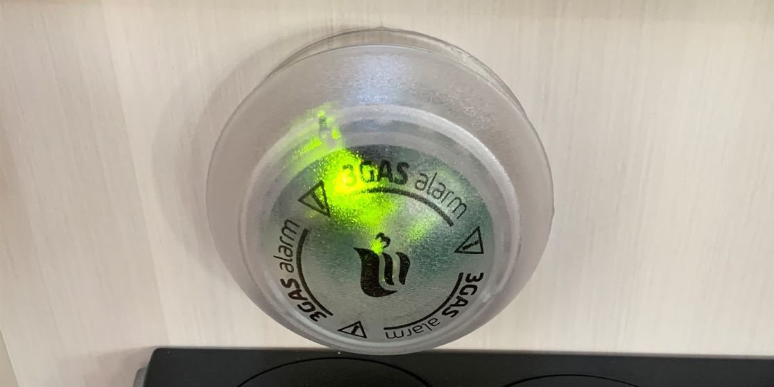 Rundes silbernes Ger&auml;t an heller Holzplatte, mittig gr&uuml;nes LED-Licht, schwarzes Flammensymbol und aufgedruckter Text 'GAS alarm', unten Teil eines schwarzen Ger&auml;ts sichtbar