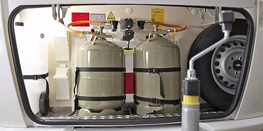 Staufach mit zwei grauen Metallgasflaschen nebeneinander, oben angeschlossene Schl&auml;uche und Regler, rechts Reserverad, vorne ein graues Rohr mit gelbem Griff