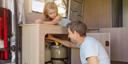 Mann kniet neben offenem Heckfach eines Wohnmobils und greift in ein Fach mit zwei Gasflaschen; ein M&auml;dchen lehnt auf der Arbeitsfl&auml;che und schaut zu; Innenraum mit Holzverkleidung sichtbar