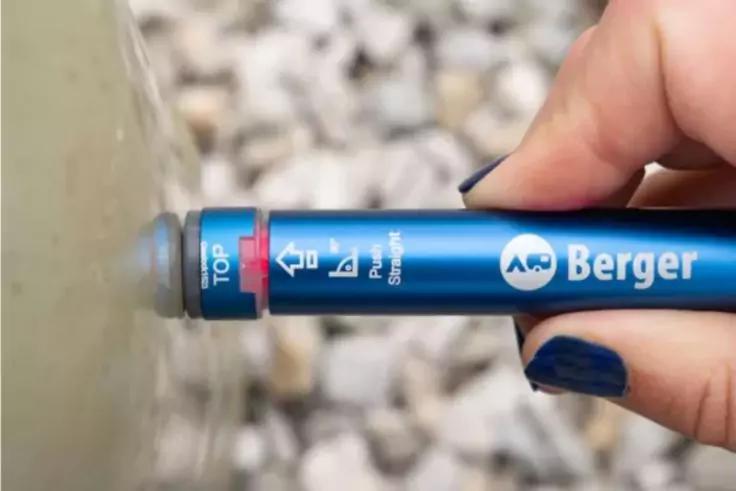 Hand mit blau lackierten N&auml;geln h&auml;lt ein blaues zylindrisches Ger&auml;t mit Schriftzug 'Berger' und Symbolen; graue Spitze ber&uuml;hrt Wasser; Kies im Hintergrund