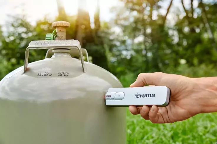 Hand h&auml;lt kleines wei&szlig;es Ger&auml;t mit Aufschrift 'truma' an eine graue Propangasflasche mit Ventil; unscharfer Hintergrund mit Gras und B&auml;umen