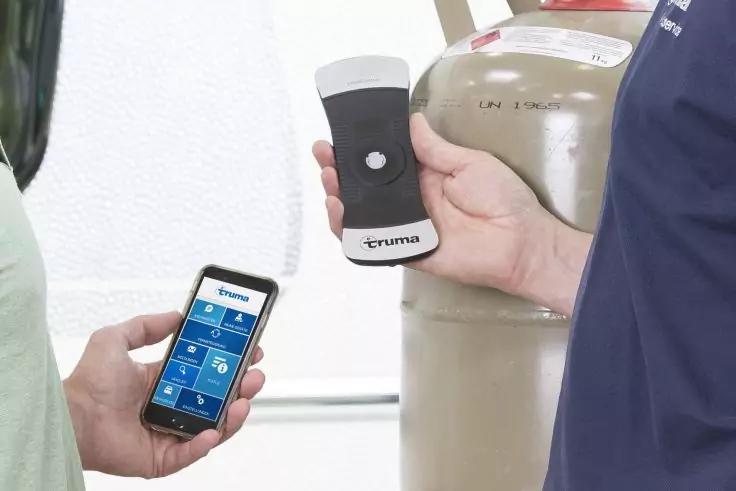 Zwei H&auml;nde: eine h&auml;lt ein Smartphone mit blauem App-Bildschirm, die andere h&auml;lt ein wei&szlig;-schwarzes Truma-Ger&auml;t vor einer Gasflasche