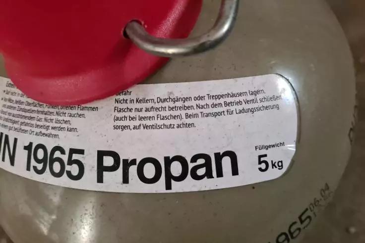 Grauer Zylinder mit wei&szlig;em Etikett 'Propan' und '5 kg', rotes Schutzteil am Ventil und Metallring sichtbar