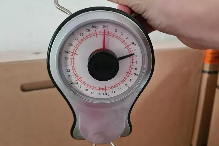 Hand h&auml;lt eine tragbare mechanische H&auml;ngewaage mit rundem Zifferblatt, roten Markierungen und schwarzer Nadel; Karton im Hintergrund