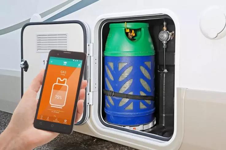 Hand h&auml;lt Smartphone mit orangefarbigem Bildschirm und wei&szlig;em Gasflaschensymbol, daneben offenes Au&szlig;enfach eines Wohnmobils mit blau-gr&uuml;ner Gasflasche