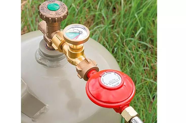 Oberseite einer grauen Gasflasche mit Manometer, Messinganschl&uuml;ssen und roter Reglerabdeckung, Gras im Hintergrund