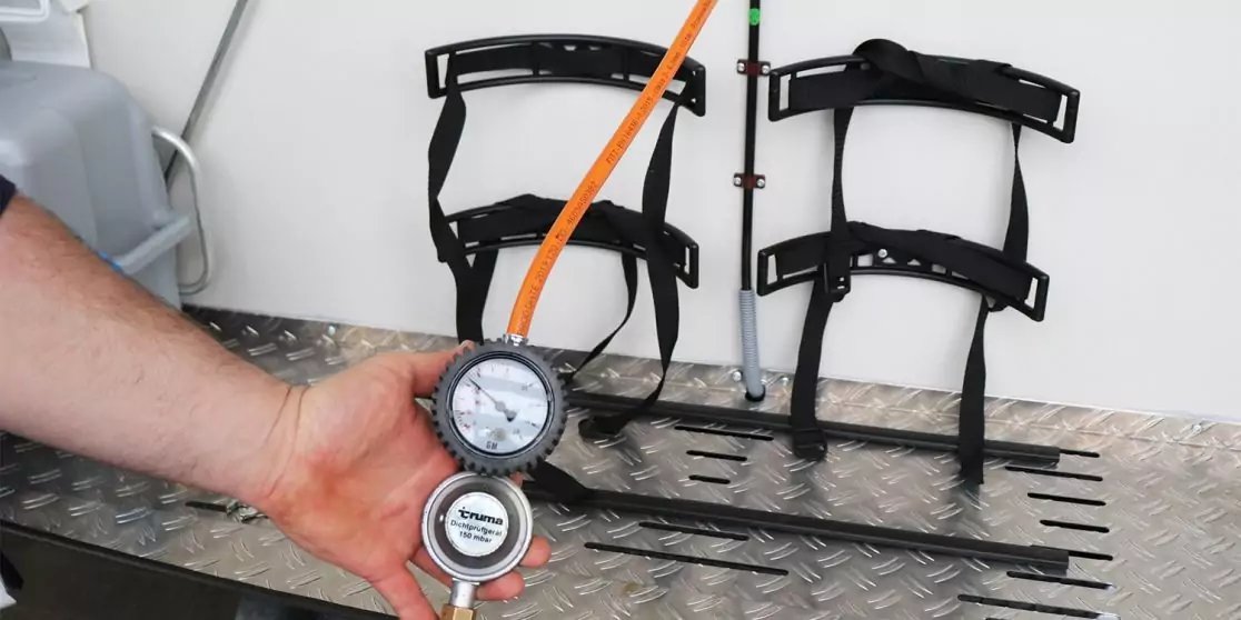 Hand h&auml;lt zwei Druckmanometer mit orangefarbener Leitung, dahinter drei schwarze Wandhalter, vorne metallische Riffelblechplatte