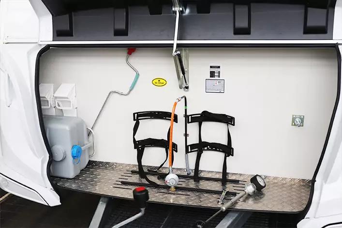 Innenraum mit wei&szlig;er R&uuml;ckwand und Riffelblechboden; links ein wei&szlig;er Kanister mit blauem Deckel; an der Wand zwei schwarze Metallgestelle mit Gurten und ein orangefarbener Gurt dazwischen; auf dem Boden ein grauer Schlauch mit silbernem Anschluss und