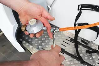 Zwei H&auml;nde halten einen silbernen Gasdruckregler; mit einer Zange wird eine orange Gasleitung am Anschluss gedreht; Riffelblech, schwarze Gurte und wei&szlig;e Verkleidung im Hintergrund