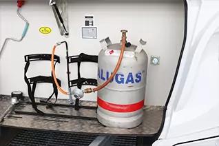 Metallgasflasche mit Ventil und Schriftzug 'ALLGAS', roter Streifen; oranger Schlauch zu schwarzen Halterungen; Metallboden und wei&szlig;e Seitenwand mit Steckdose und gelbem Smiley-Aufkleber