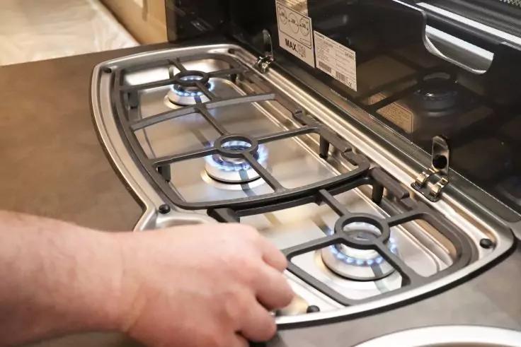 Hand an Drehknopf eines Gaskochfelds, drei sichtbare blaue Flammen