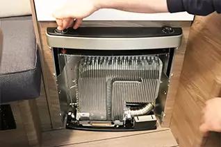 Ge&ouml;ffnete Frontabdeckung eines Ger&auml;ts mit sichtbaren metallischen Lamellen, Rohren und Kabeln; zwei H&auml;nde halten die Abdeckung; Holzfu&szlig;boden und Sofa daneben