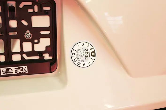 Nahaufnahme der Fahrzeugkarosserie mit links einem schwarzen Kennzeichenhalter und rechts einer runden Plakette mit der Jahreszahl 2022 auf wei&szlig;em Lack