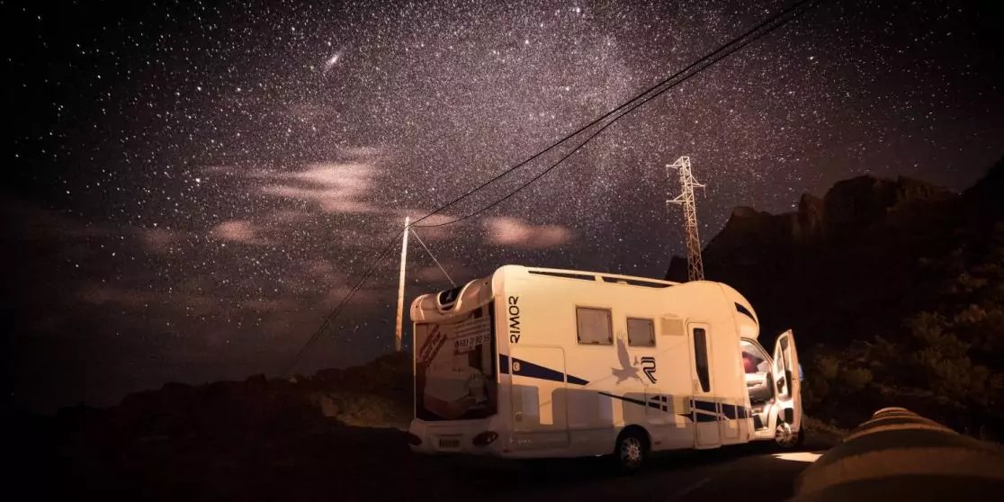 Wohnmobil auf dunkler Landstra&szlig;e neben Felsen und einem Strommast mit Leitungen, dar&uuml;ber dichter Sternenhimmel mit sichtbarer Sternenband