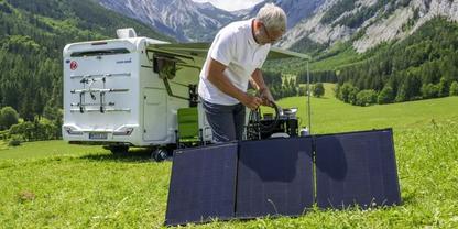 Ein Mann mit grauen Haaren steht an einem aufgestellten faltbaren Solarpanel auf einer Wiese neben einem wei&szlig;en Wohnmobil, Berge im Hintergrund