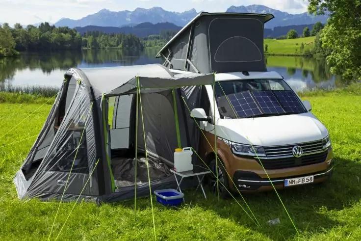 Campingbus mit aufgeklapptem Schlafdach, angebautem Vorzelt und Solarpanel, auf einer Wiese neben einem See mit Bergen im Hintergrund