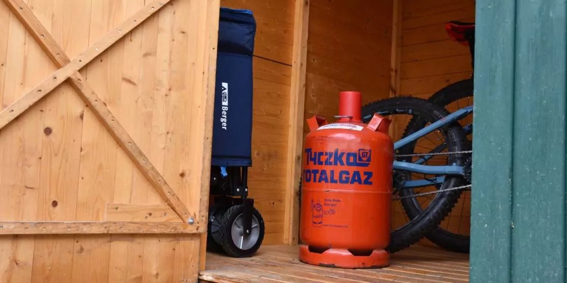 Innenraum eines Holzschuppens mit ge&ouml;ffneter T&uuml;r, im Vordergrund eine orange Gasflasche mit Aufschrift, daneben ein gefaltetes Objekt mit blauem Bezug und R&auml;dern sowie ein Fahrradreifen