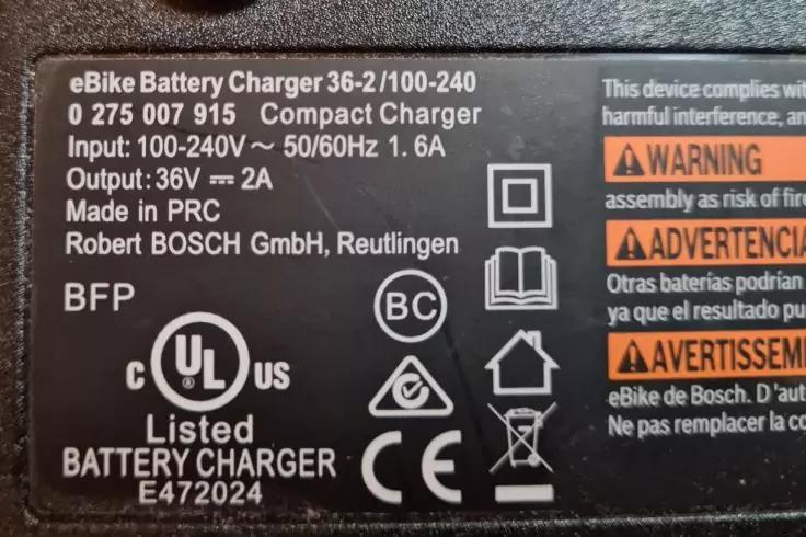 Nahaufnahme eines schwarzen Etiketts mit weißer Schrift: 'eBike Battery Charger', Modellnummern, Eingangs- und Ausgangswerte, 'Robert BOSCH GmbH, Reutlingen', mehrere Sicherheitssymbole und orange Warnfelder