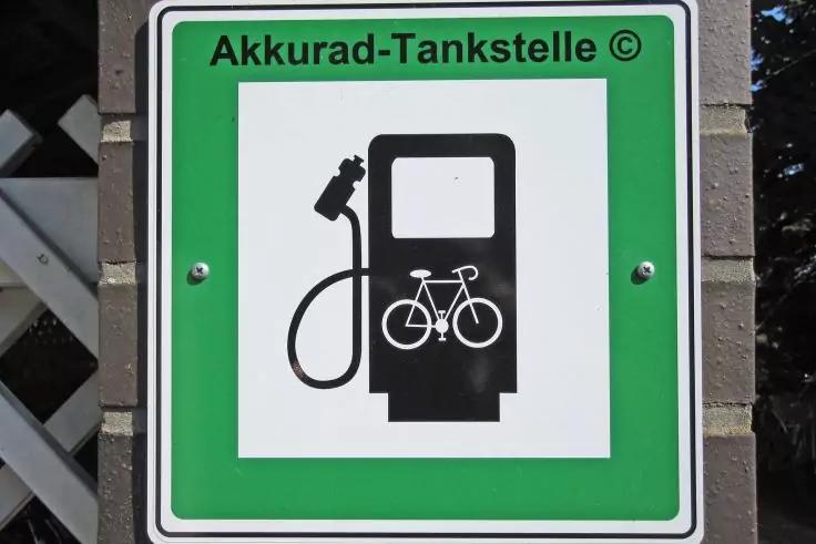 Grünes Schild mit weißem Quadrat, schwarze Zapfsäule mit Fahrrad-Symbol und Zapfpistole; oben Schrift 'Akkurad-Tankstelle ©'