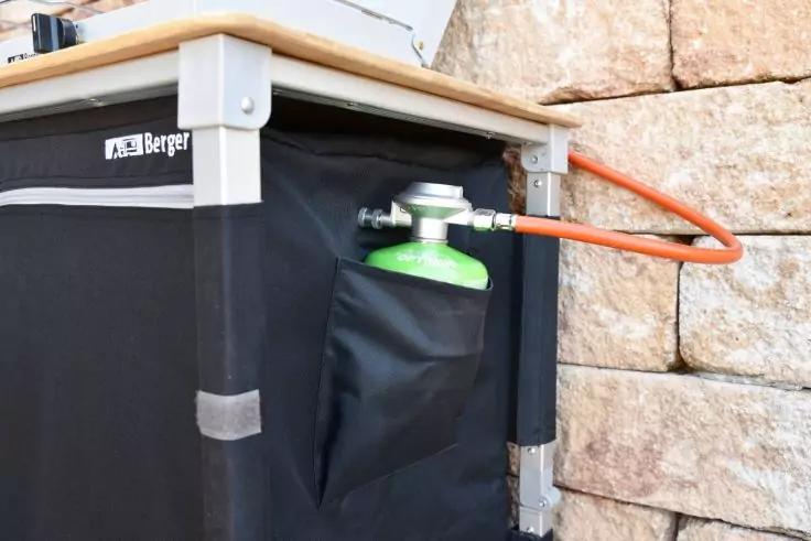 Schwarzer Unterschrank mit Holzplatte, an der Seite eine gr&uuml;ne Gaspatrone mit Metallregler und orangefarbenem Schlauch, im Hintergrund eine Steinmwand