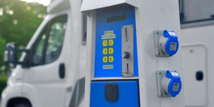 Blaue S&auml;ule mit gelbem Tastenfeld und zwei blauen runden Steckdosen, im Hintergrund ein wei&szlig;es Wohnmobil und unscharfe B&auml;ume