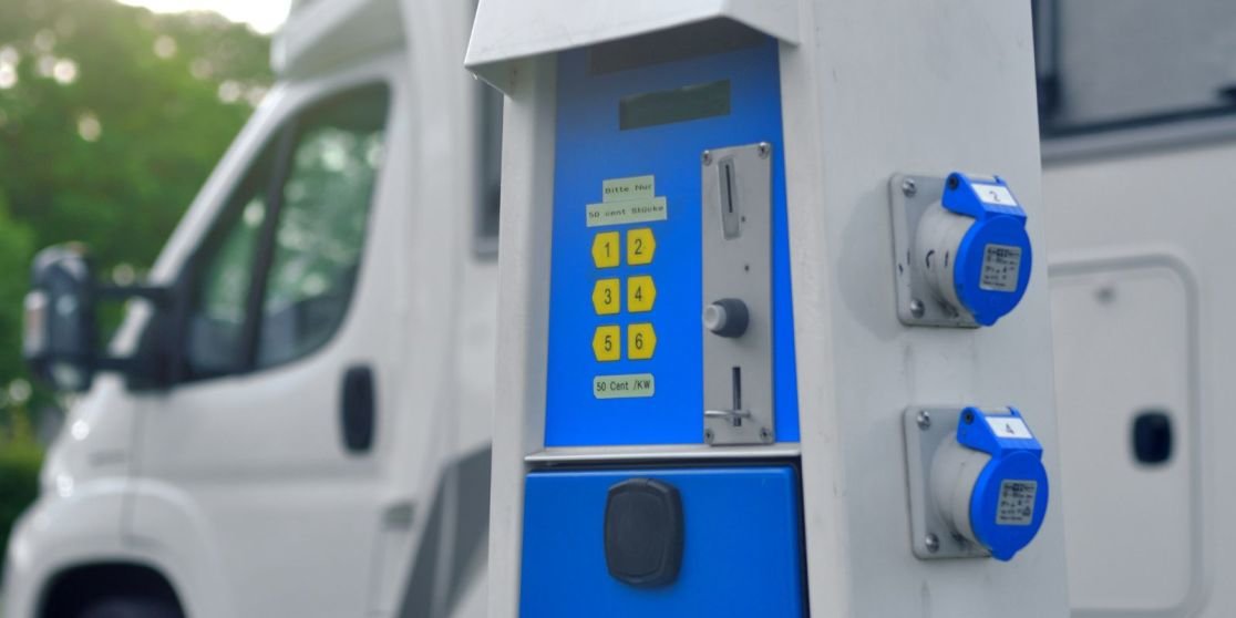 Blaue S&auml;ule mit gelbem Tastenfeld und zwei blauen runden Steckdosen, im Hintergrund ein wei&szlig;es Wohnmobil und unscharfe B&auml;ume