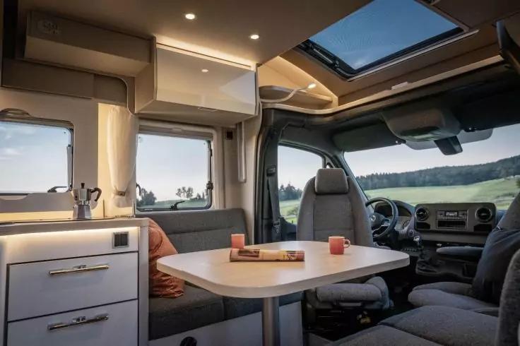 Innenraum eines Wohnmobils mit Tisch und Sitzbank, Einbauk&uuml;che mit Sp&uuml;le und Schubladen, Oberschrank, zwei Fenster, Dachfenster und Fahrer- sowie Beifahrersitz
