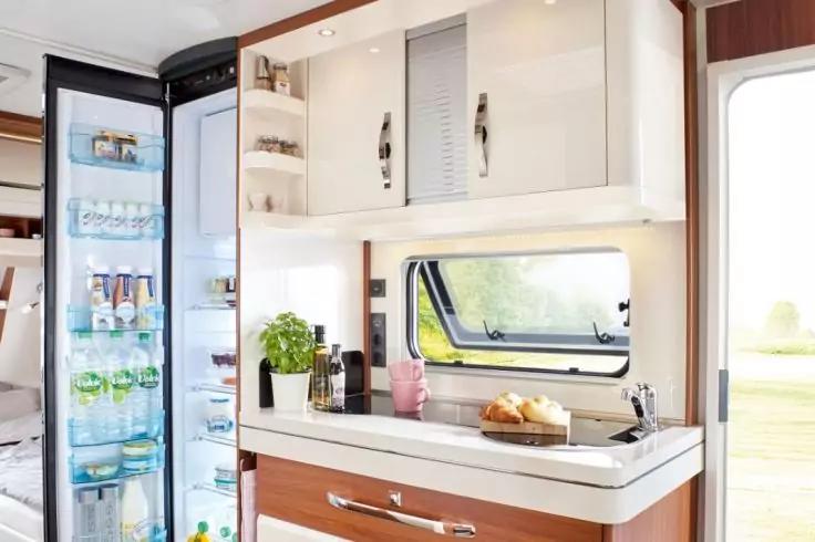 K&uuml;chenzeile in einem Fahrzeug mit ge&ouml;ffnetem K&uuml;hlschrank, Arbeitsfl&auml;che mit Topfpflanze, Schneidebrett mit Br&ouml;tchen, Sp&uuml;le, Oberschr&auml;nken und Fenster