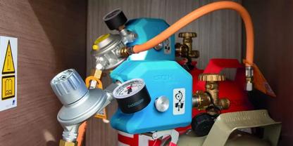 Blaues Ger&auml;t mit Manometer, orange Schlauch, roter Metallzylinder, Messingarmaturen, innen in einem Holzschrank, wei&szlig;er Warnaufkleber mit gelbem Dreieck