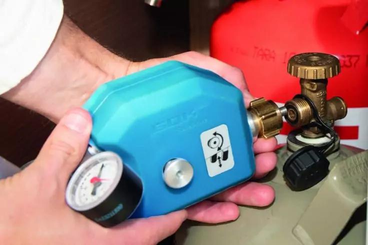 Hände halten ein blaues Gerät mit Manometer an einem Messingventil einer roten Gasflasche