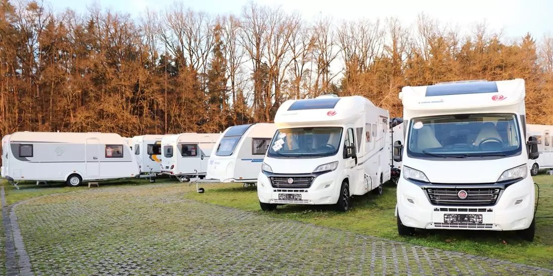 Mehrere wei&szlig;e Wohnmobile auf einem gepflasterten Stellplatz mit grasbewachsenen Gittersteinen, im Hintergrund kahle B&auml;ume, keine Personen