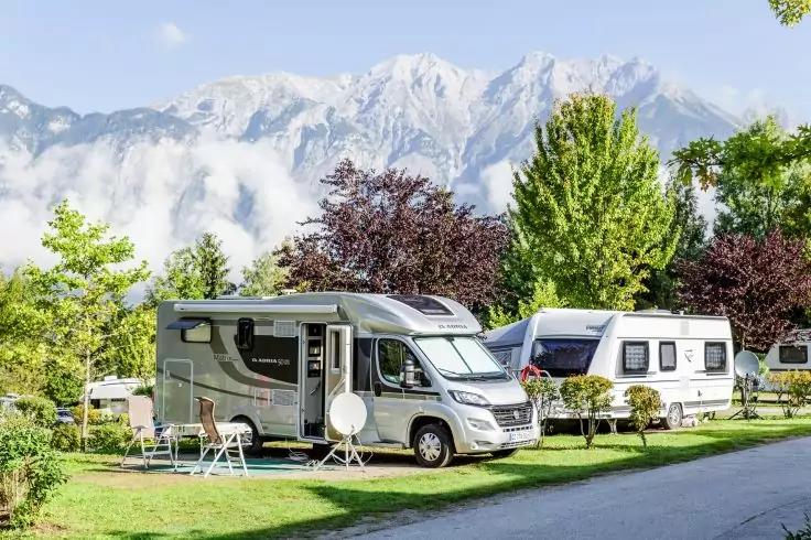 Wohnmobil vorn und Wohnwagen dahinter auf einem grasbewachsenen Campingplatz, Klapptisch und St&uuml;hle neben dem Wohnmobil, B&auml;ume und Berge im Hintergrund, Weg im Vordergrund