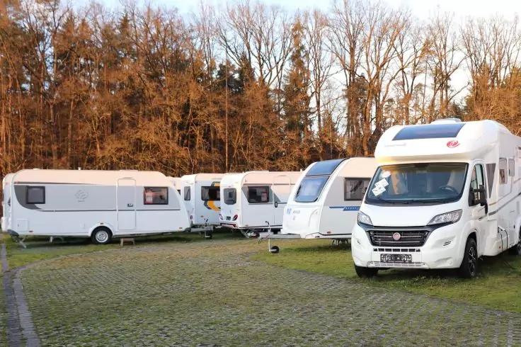Mehrere Wohnwagen und ein Wohnmobil auf einer gepflasterten, teils grasbewachsenen Fl&auml;che, im Hintergrund eine Baumreihe