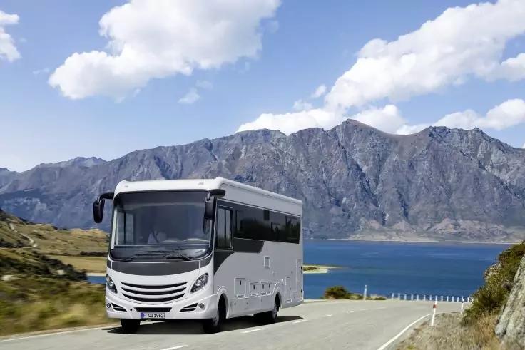 Wei&szlig;es Wohnmobil auf K&uuml;stenstra&szlig;e, Wasserfl&auml;che, Bergkette und wolkiger Himmel im Hintergrund