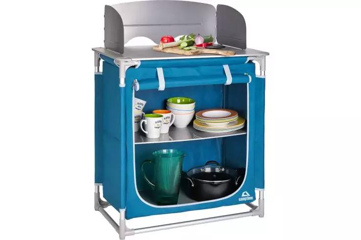 Klappbarer Schrank mit blauem Stoffbezug und Aluminiumgestell, zwei Regalböden mit Tassen, Schalen und Tellern; darunter grüne Kanne und schwarzer Topf; oben Schneidebrett mit geschnittenem Gemüse und grauem Windschutz