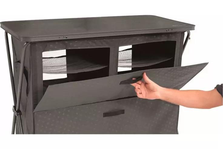 Graue Stoff-Aufbewahrung mit flachem Deckel, zwei rechteckige offene Fächer oben, vordere Klappe wird von einer rechten Hand geöffnet, sichtbares Metallgestänge an den Seiten, weißer Hintergrund